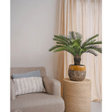 Een Kunstplant Cycaspalm 80 cm staat op een ronde rieten tafel naast een beige fauteuil met een wit kussen met patroon, tegen lichtbeige gordijnen en een neutrale muur.
