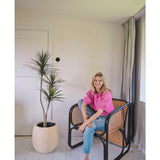 Een vrouw in een felroze topje en blauwe spijkerbroek zit op blote voeten op een moderne zwarte en rieten stoel naast de Kunstplant Dracaena Fragnans 150 cm in een lichte, minimalistische kamer met witte muren en vloer.