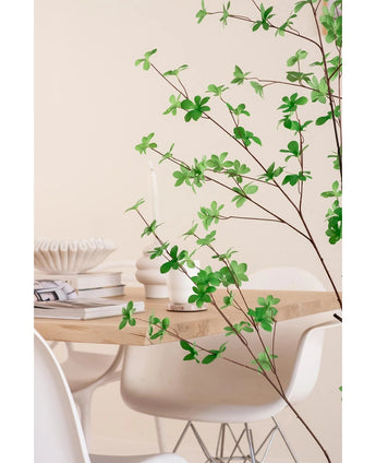 Een moderne eethoek met witte stoelen en een lichthouten tafel, gedecoreerd met boeken, kaarsen en een witte vaas, wordt op de voorgrond omlijst door de Kunstplant Enkianthus 150 cm bladgroene plant.