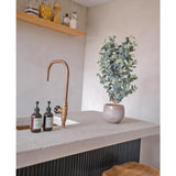Een modern aanrecht heeft een messing kraan, zeepdispensers, houten krukjes en de Kunstplant Eucalyptus 91 cm. Glazen potten met voorraadartikelen staan op een houten plank tegen een lichtgekleurde muur.