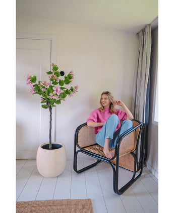 Een vrouw in een roze trui en blauwe spijkerbroek zit op blote voeten op een rotan stoel naast de Kunstplant Judas 150 cm, een kunstboom met roze bloemen, in een lichte, moderne kamer met witte muren en vloeren.