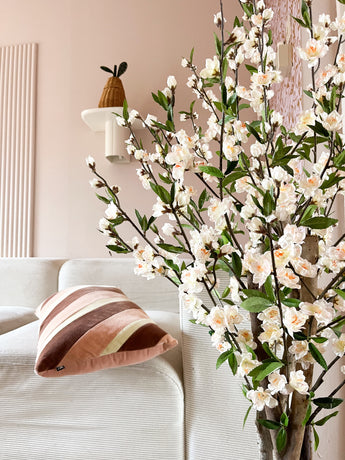 Een gezellige woonkamer met een witte bank, een gestreept roze en bruin kussen en de Kunstplant Kersenbloesem 120 cm crème bloeiende tak op de voorgrond. Een potplant rust op een wandplank tegen een lichtroze muur.