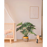 Een gezellige, minimalistische kinderkamer met links een houten wieg en luifel, in het midden een Kunstplant Philodendron UV 90 cm als pronkstuk, rechts een houten speelgoedfiets en daarboven een eenvoudige lijnkunstprint.