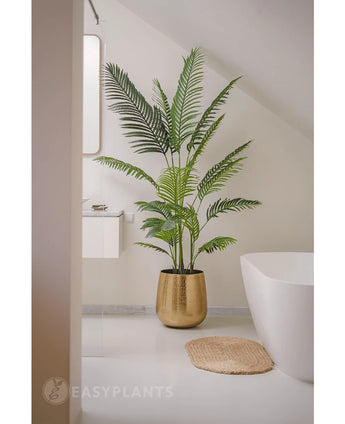 De Kunstplant Phoenix 180 cm in een gouden pot staat bij een wit bad en wastafel in een moderne minimalistische badkamer met witte muren en vloer. Vlakbij ligt een ronde geweven mat op de vloer.