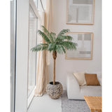 De Kunstplant Phoenix Palm De Luxe 180 cm in een pot met structuur staat bij grote ramen in een lichte woonkamer met neutrale inrichting, met een witte bank, beige kussens, een houten tafel en abstracte kunst aan de muur.