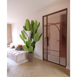 Een moderne woonkamer met een witte bank, gedessineerde kussens en de Kunstplant Strelitzia Real Touch Deluxe 210 cm. Een lichthouten vloer, zachtroze muren en een glazen deur met een bronzen metalen frame creëren een lichte, luchtige sfeer.