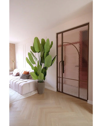 Een moderne woonkamer met een witte bank, gedessineerde kussens en de Kunstplant Strelitzia Real Touch Deluxe 210 cm. Een lichthouten vloer, zachtroze muren en een glazen deur met een bronzen metalen frame creëren een lichte, luchtige sfeer.