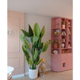 De Kunstplant Strelitzia Real Touch Deluxe 210 cm staat bij een cheetafiguur van keramiek met een oranje hoed, naast een hoge roze plank vol vazen, bloemen en ornamenten in een pastelkleurige kamer.