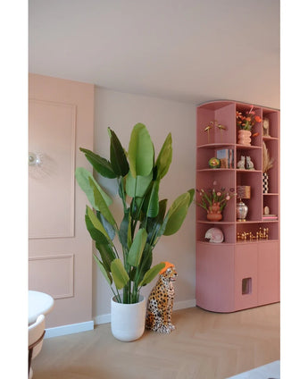 De Kunstplant Strelitzia Real Touch Deluxe 210 cm staat bij een cheetafiguur van keramiek met een oranje hoed, naast een hoge roze plank vol vazen, bloemen en ornamenten in een pastelkleurige kamer.