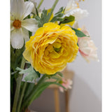 Een close-up van de Kunstranonkel Real Touch Geel 60cm, een levendige gele kunstranonkel, omringd door witte bloemen en groene bladeren in een vaas binnenshuis, met een zacht onscherpe achtergrond.