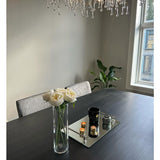 Een moderne eettafel met de Kunstrozenboeket "Carmen" Real Touch Crème wit 36cm als middelpunt, een spiegelplateau met kaarsen en decor, een grijs gestoffeerde stoel, een bladplant bij het raam en een kristallen kroonluchter boven.