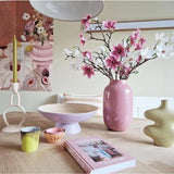 Op een lichte houten tafel staat de Kunsttak Magnolia kleinbloemig 89 cm wit in een roze vaas, modern keramiek, kleurrijke kopjes, een schaal, een gele kaars in een unieke houder en een roze boek, met bloemenkunstwerk op de muur erachter.