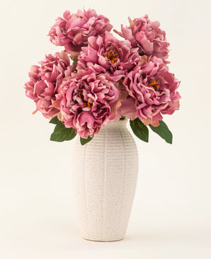 De Zijden kunstbloemen boeket Peony Dusty Pink is gevat in een witte vaas van keramiek met textuur. De vaas heeft grote roze pioenrozen met weelderige groene bladeren tegen een lichte, effen achtergrond.