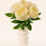 Een golvende, gestructureerde witte vaas bevat het Zijden kunstbloemen boeket Peony Wit-grote crèmekleurige pioen kunstbloemen met groene bladeren tegen een effen lichte achtergrond.