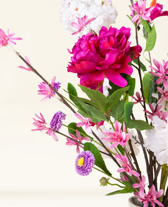 Het Zijden kunstbloemen boeket Pink Reverie heeft een grote magenta pioenroos, kleine roze bloemen, paarse madeliefachtige bloemen en groene bladeren in een vaas-een romantisch boeket tegen een lichte achtergrond.