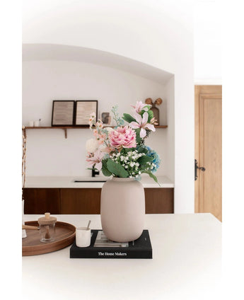 De Vaas Artic Beige Ø21 x H29 cm, gevuld met roze en blauwe kunstbloemen, staat op een zwart boek op een wit aanrecht. Zachte verlichting accentueert het minimalistische tafereel met decorplanken en een houten deur op de achtergrond.