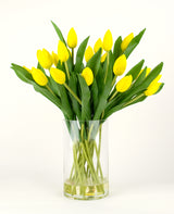 Het Zijden kunstboeket Tulpen in vaas Geel bestaat uit een boeket gele tulpen met groene bladeren in een heldere glazen vaas, tegen een effen witte achtergrond.
