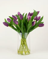 Het Zijden kunstboeket Tulpen in vaas Paars heeft langstelige paarse kunsttulpen met weelderige groene bladeren, prachtig gerangschikt in een heldere glazen vaas tegen een effen witte achtergrond.