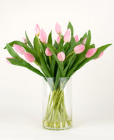 Het Zijden kunstboeket Tulpen in vaas Roze heeft roze tulpen met lange stelen en groene bladeren in een heldere glazen vaas, tegen een effen witte achtergrond.