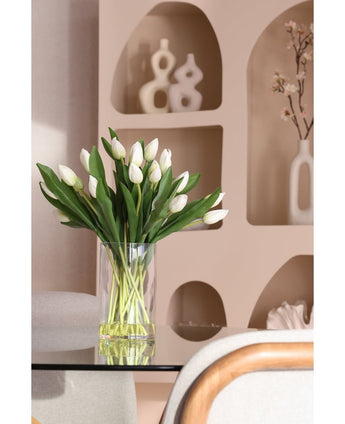 Het Zijden kunstboeket Tulpen in vaas Wit, met witte tulpen en groene bladeren, staat op een glazen tafel. Daarachter staan op beige planken moderne keramische decorstukken en een bloeiende tak in een witte vaas.